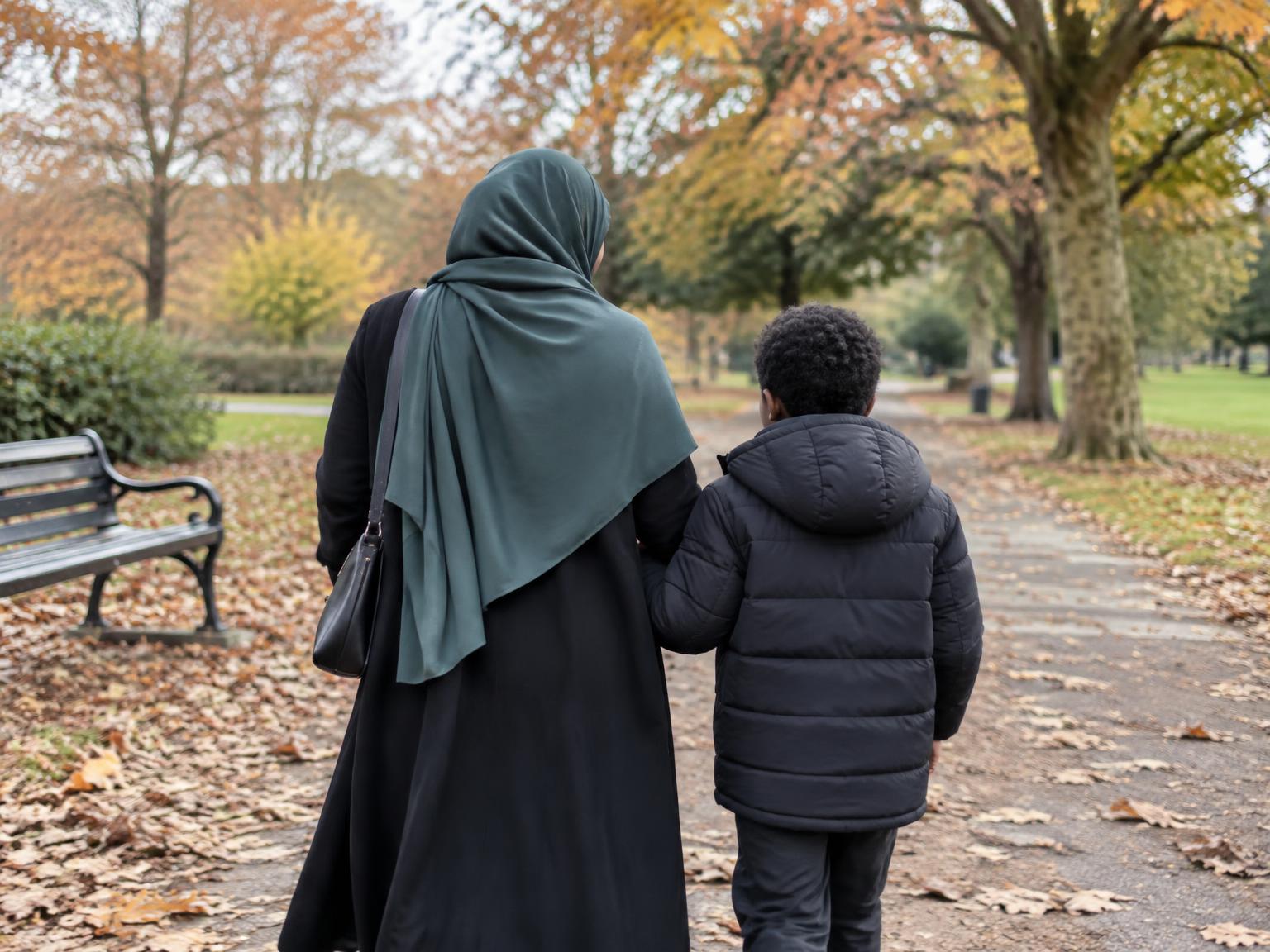 A Muslim mother and son walking together through a park, seen from behind